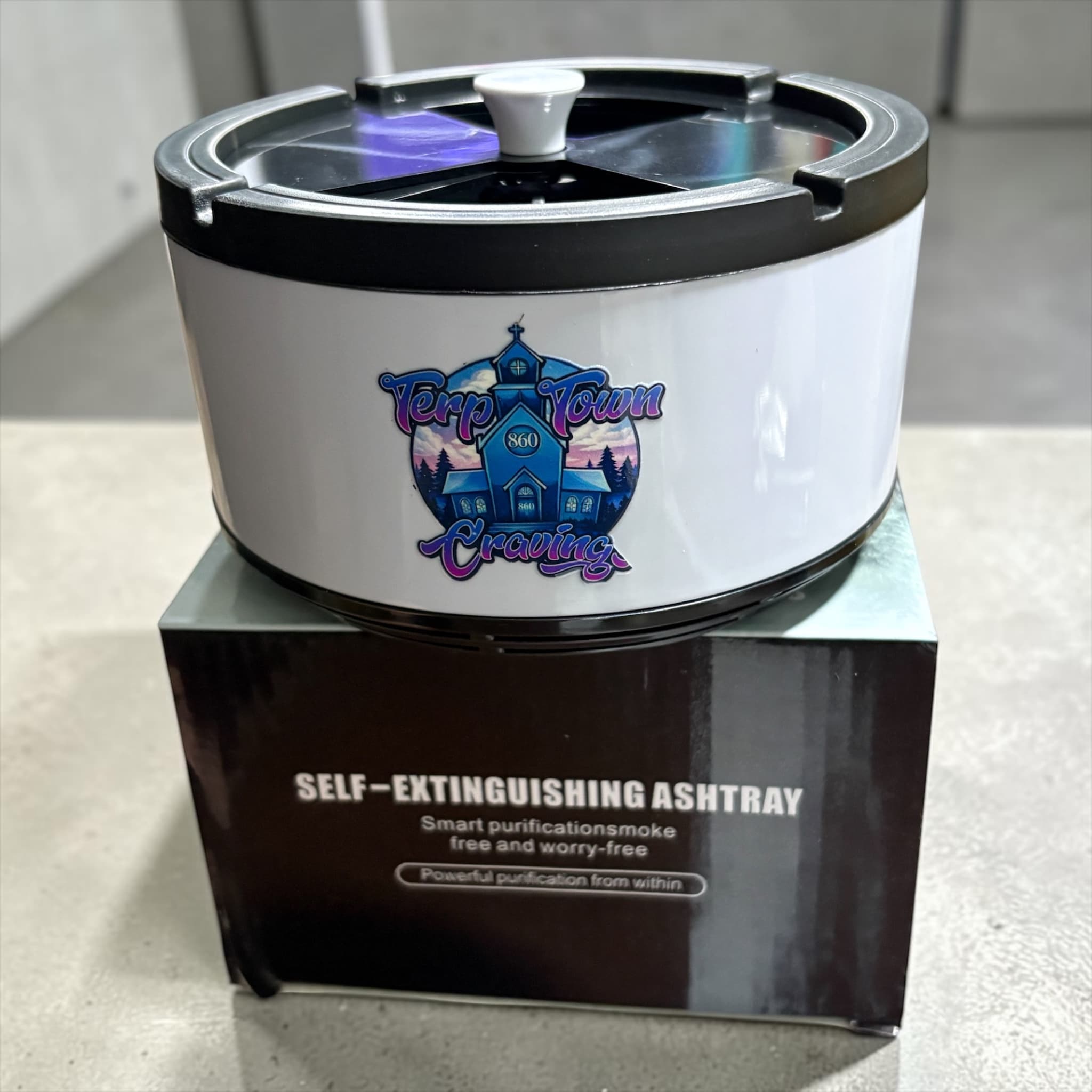 White self-extinguishing ashtray with a colorful logo sitting on top of its black product box.