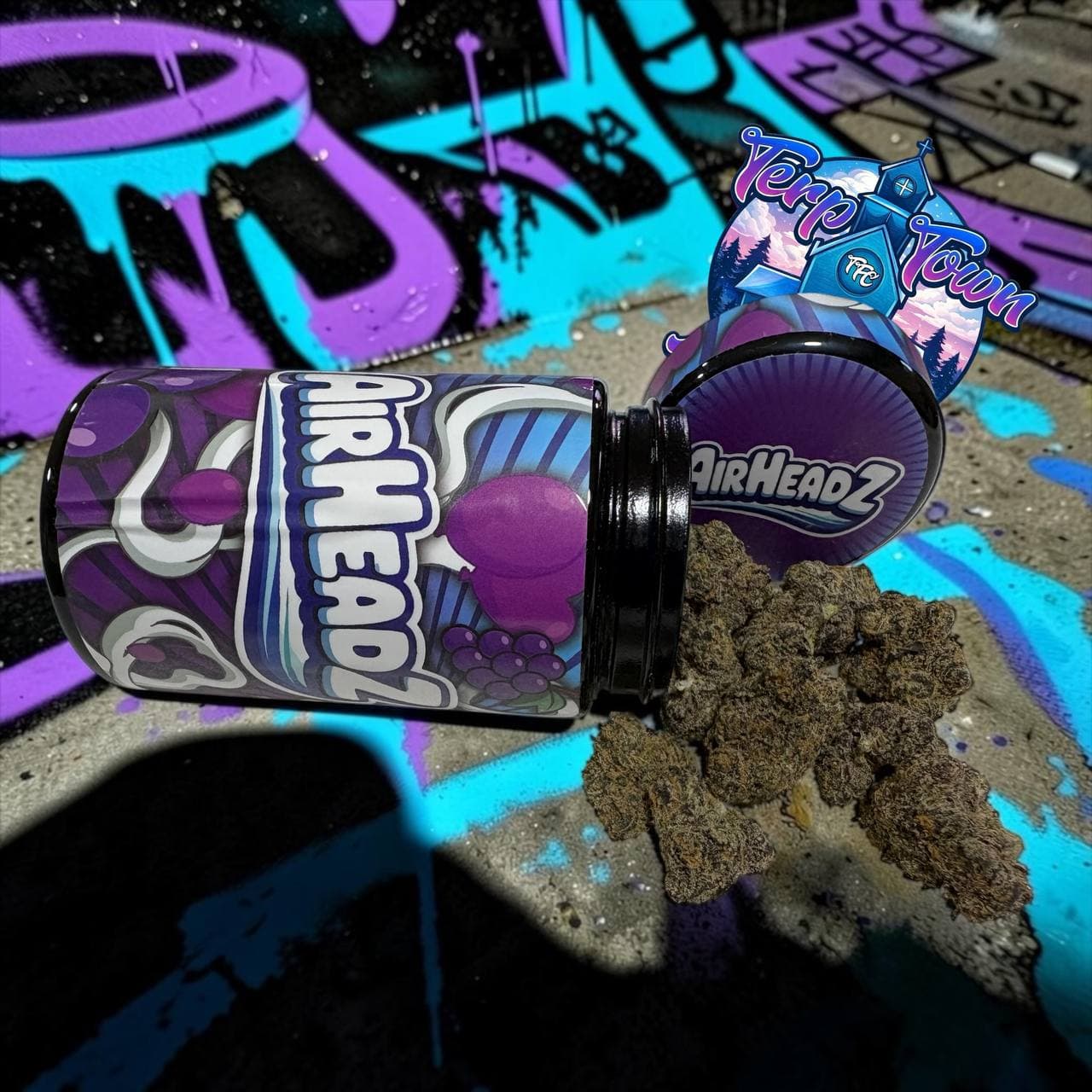 Airheadz cannabis jar spilling dark, dense buds onto a concrete surface with purple graffiti.