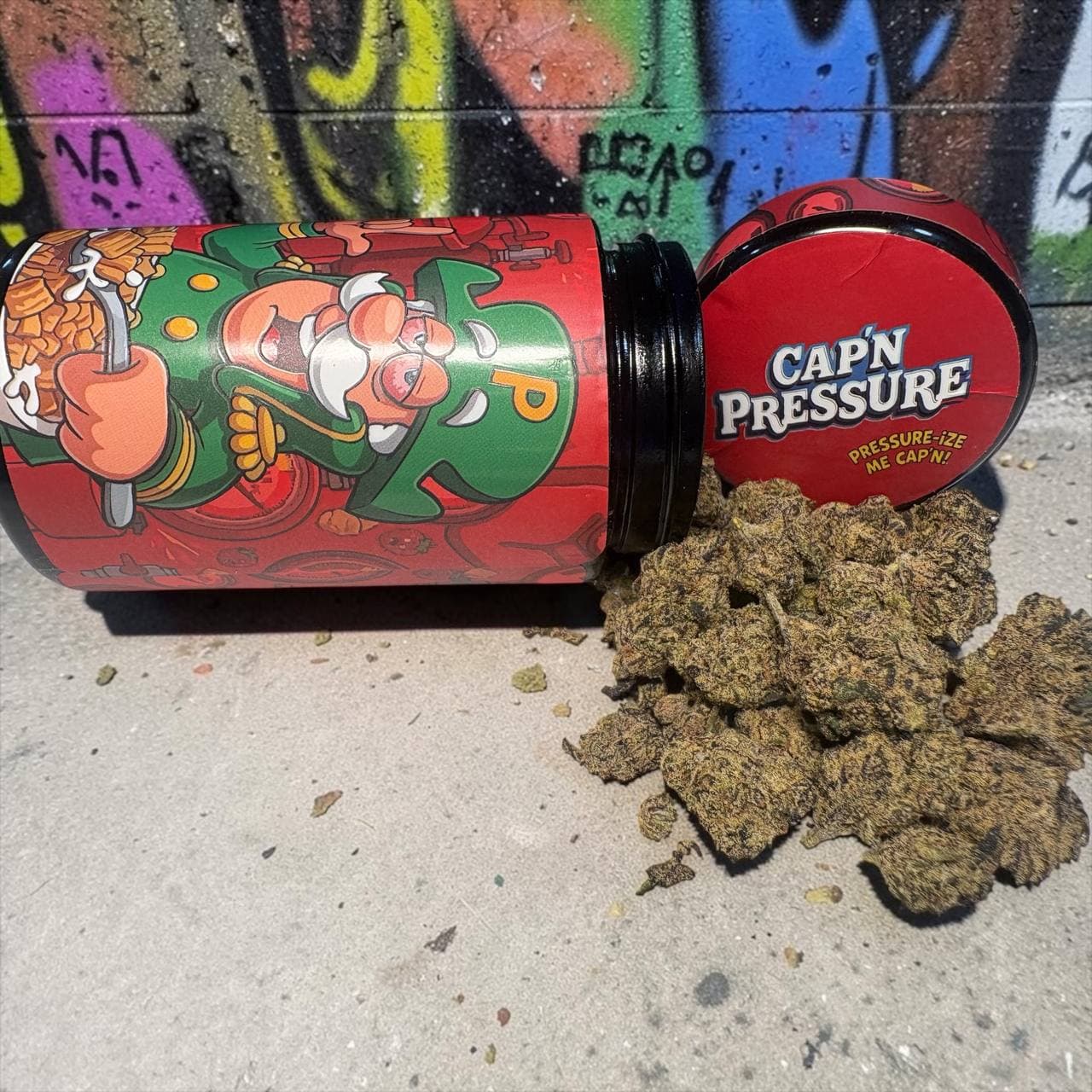 Dark cannabis buds spilled from a red Cap'n Pressure jar onto concrete against graffiti.